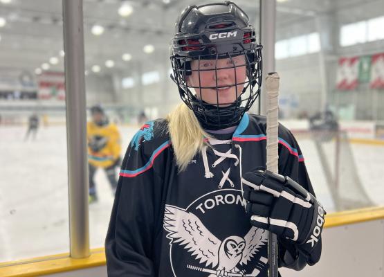Girl with blonde hair in hockey gear holding stick with people on rink behind her