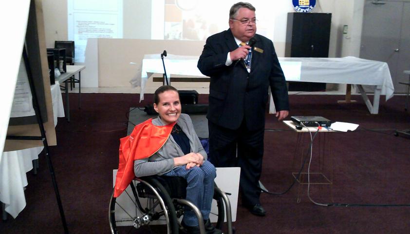 Two people in office. Woman on left uses a wheelchair and is wearing a cape, person on the right is in a suit.