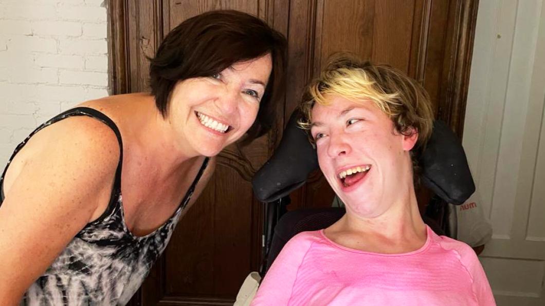 Two women, one in a black and white shirt and the other in a pink shirt with wheelchair headrest, lean in and smile