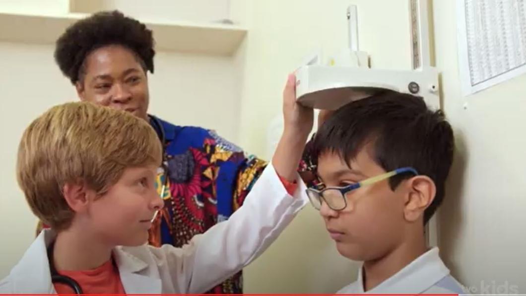 A child pretending as doctor to check the height of another child, an adult is standing behind them.