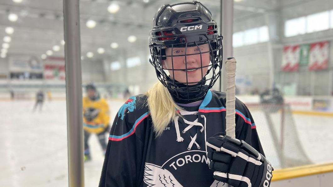 Girl with blonde hair in hockey gear holding stick with people on rink behind her
