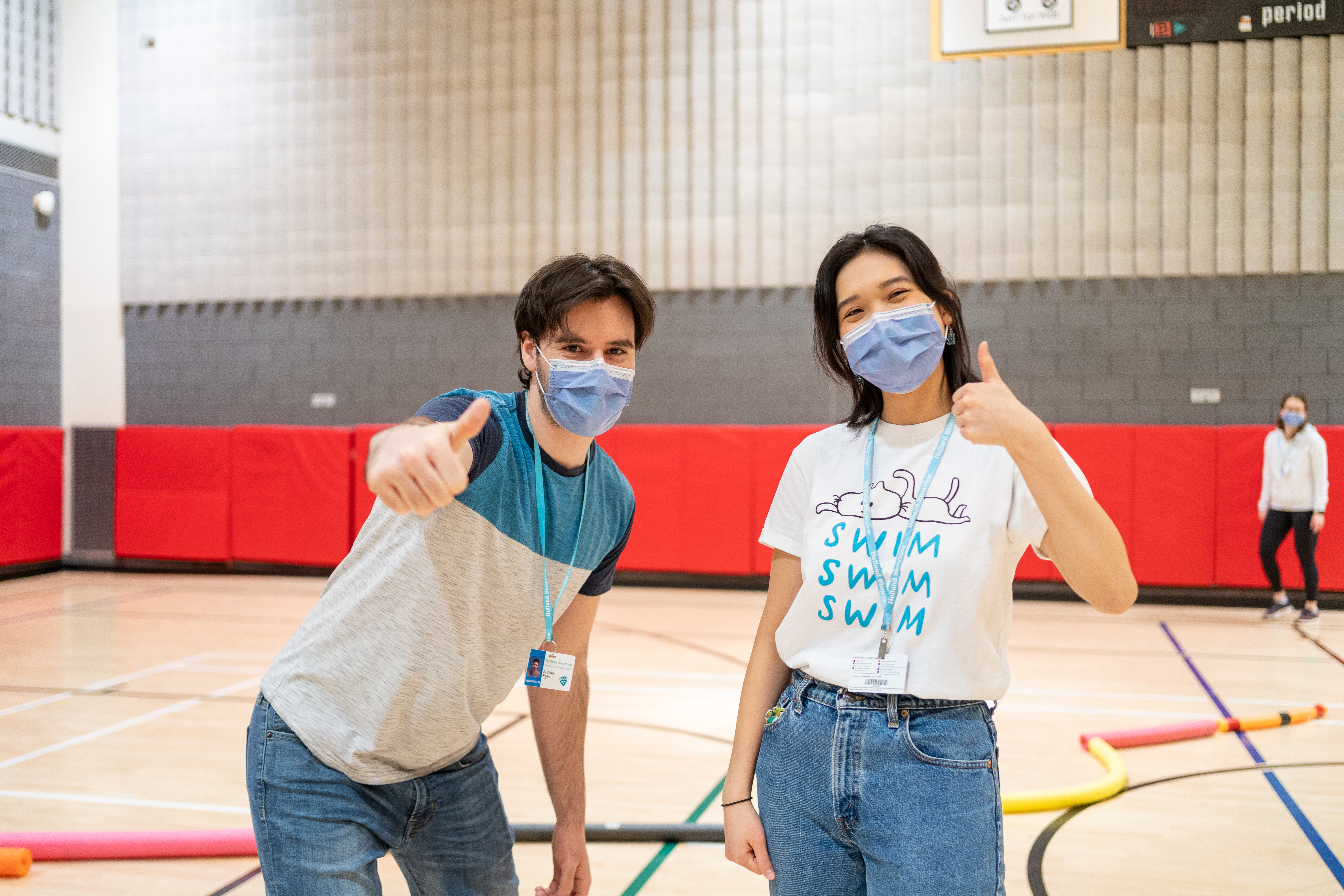 Two volunteers stand masked in a gymnasium giving a thumbs up at the camera.