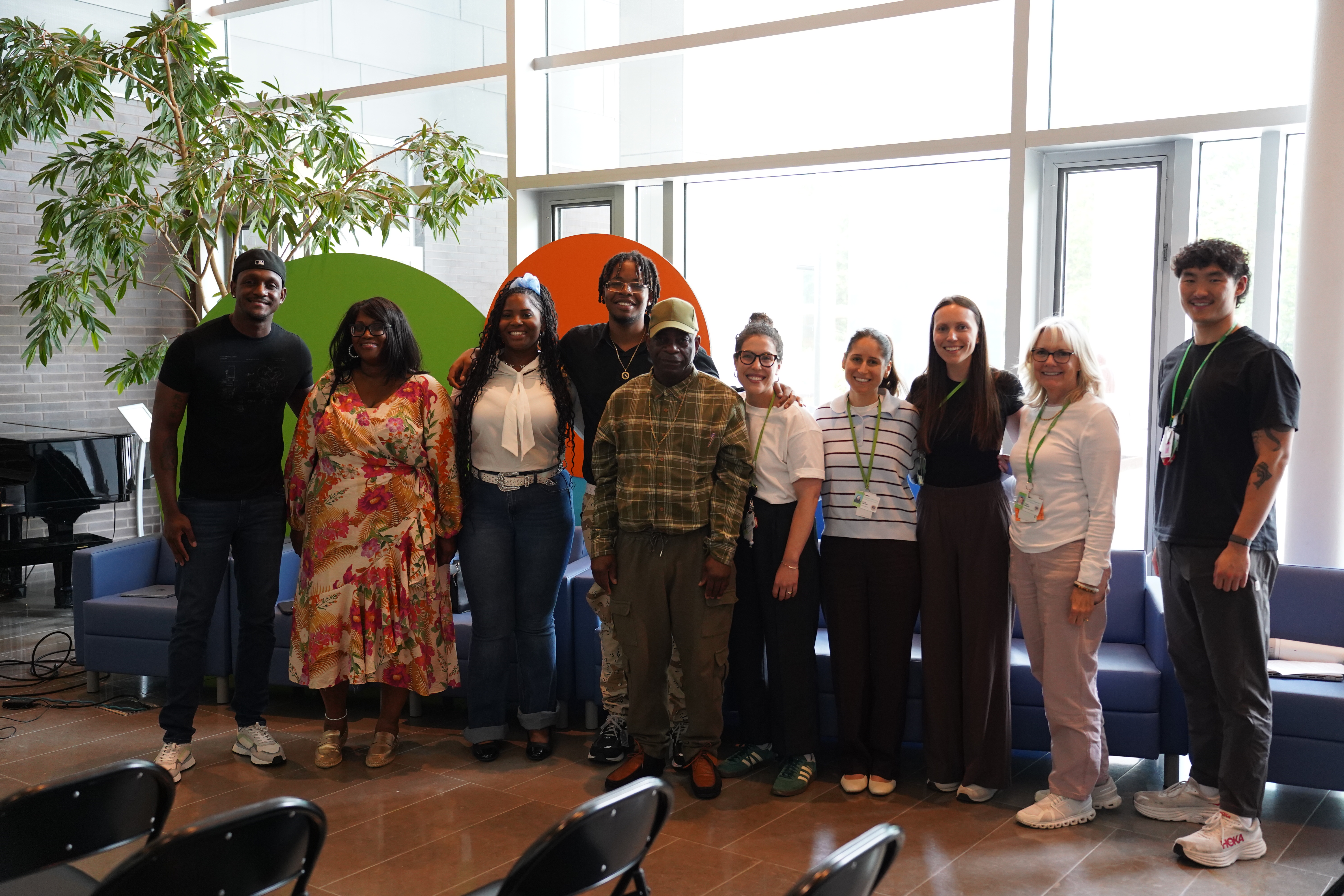 Photograph of a diverse group of ten people standing indoors in front of large windows and colorful circular wall decorations. They are wearing varied attire including dresses, casual wear, and lanyards