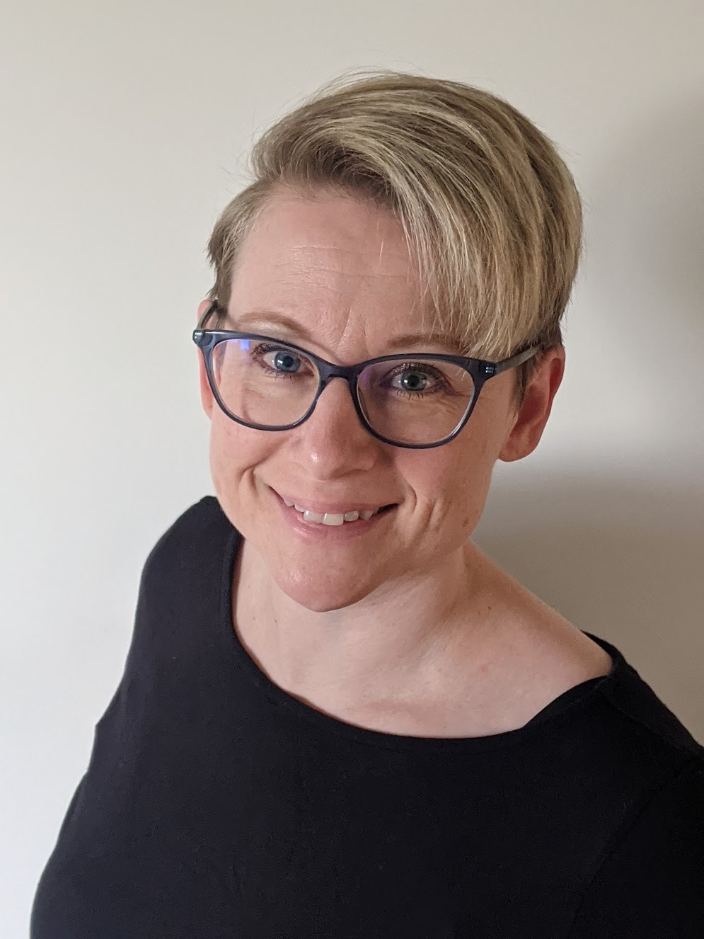 Caucasian female with short blonde hair, black shirt and black rimmed glasses
