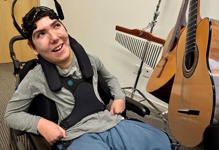 A teenager in a wheelchair wearing a BCI headset and playing a guitar