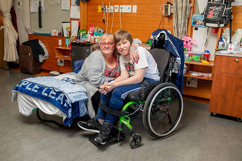 A teenager sitting on a wheelchair is being hugged by a senior, who is sitting on a hospital bed