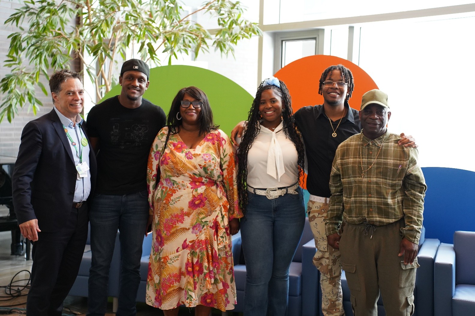 Photograph of six people standing indoors in front of colorful circular wall decorations in green, orange, and blue. Group includes individuals wearing casual and business attire, with a plant visible on the left and seating furniture in the background.