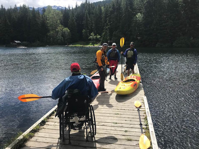 3 men standing on a dock with kayaks and a man sitting in a wheelchair holding a paddle