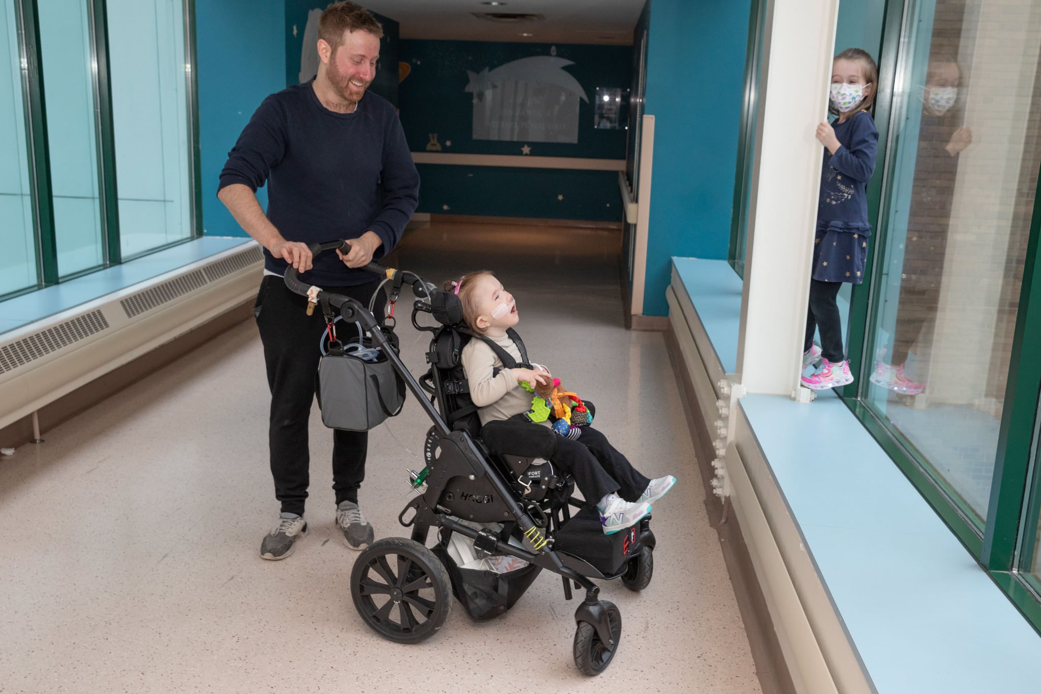 Man with beard pushes girl in stroller while another girl hides behind a barrier