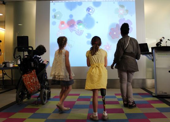 Children, two with disabilities, stand on coloured tiles in front of a screen with images on it.