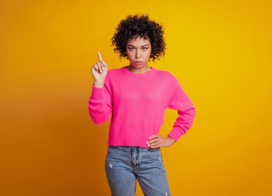 Woman with curly hair and pink shirt, one hand on hip and the other pointing in the air