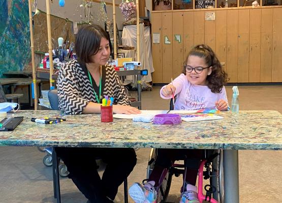 Child with dark hair in pink wheelchair sits at table painting with a woman beside her in an art studio