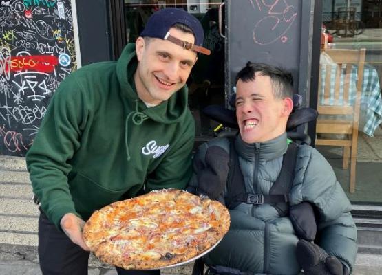 Young man with dark hair grins in wheelchair beside man with basketball cap holding a pizza