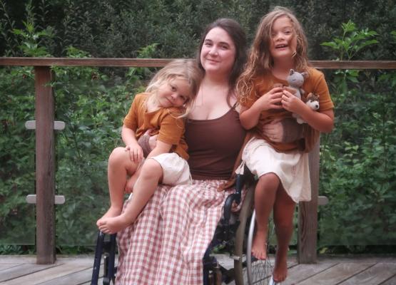 Woman with long dark hair in wheelchair with a young girl snuggled on her lap and older girl balanced on the side