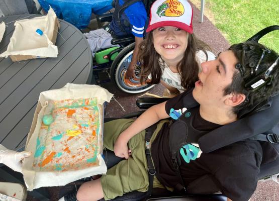 Boy in wheelchair wearing head set sits in front of a paint ball activity while girl in baseball cap smiles