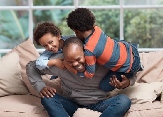 Man on couch laughs with two young children hanging off his neck.