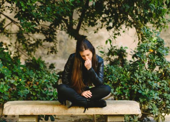 Young woman with long dark hair sits on bench with head down in her hand