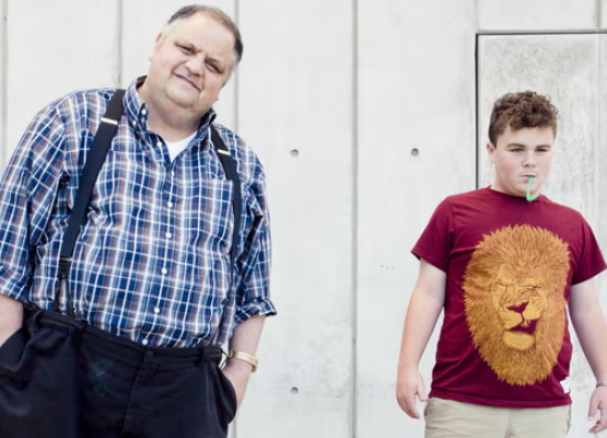 Man in plaid shirt with suspenders stands beside teen with red shirt with gold lion on it