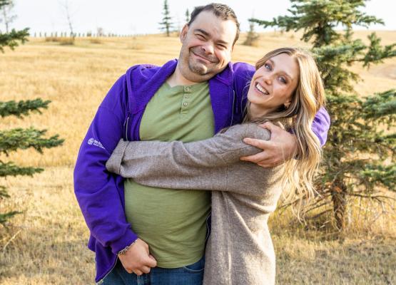 Woman with long light brown hair wraps arms around man wearing a green t-shirt and purple sweatshirt