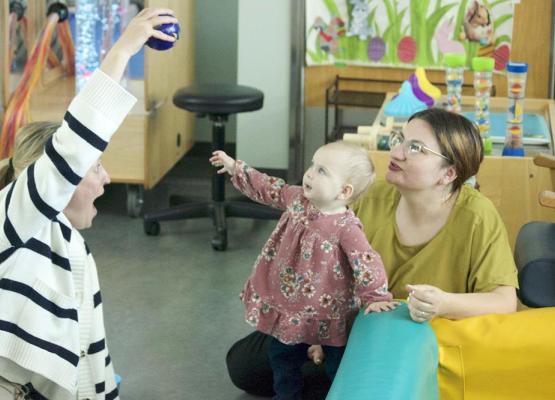 Toddler in pink dress reaches up to a ball held by a woman in a striped sweater while another woman smiles