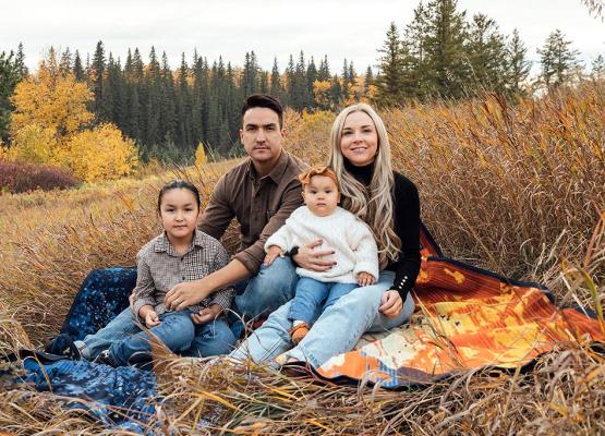 Man with dark hair beside woman with long blonde hair with boy and baby girl in front on a blanket in a field