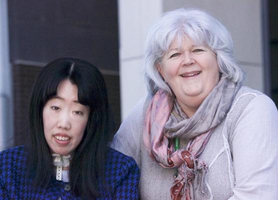 Woman with long black hair in navy sweater with a tracheotomy cork in her neck smiles with woman with grey hair wearing a pink and purple scarf