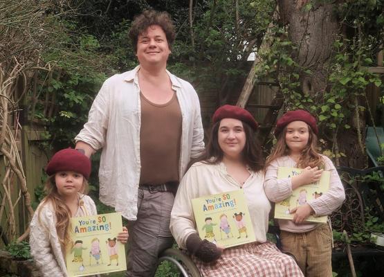 A man with brown hair stands behind a woman in a wheelchair and two girls who are each holding a children's book