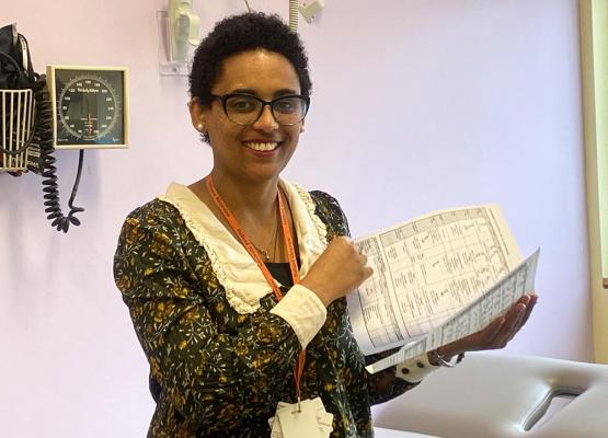 Woman with dark hair and medical equipment behind her holds a document and smiles