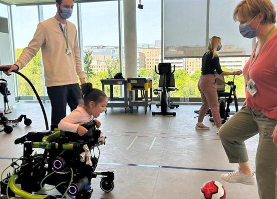 Small girl in green robot-powered walker faces therapist who is kicking a soccer ball