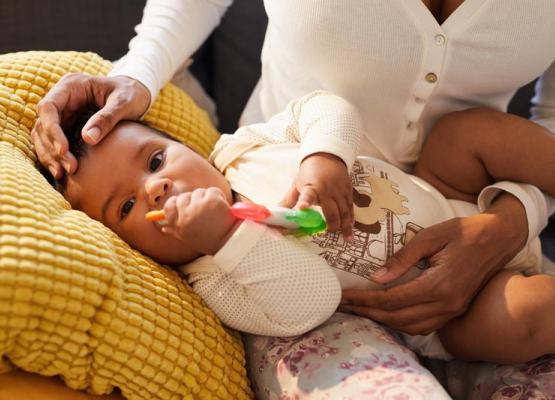 A baby holds an orange and green toy on a pillow balanced on mother's lap