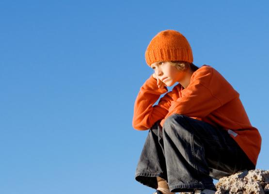 Child in orange sweater and hat sits on rock with head on fist