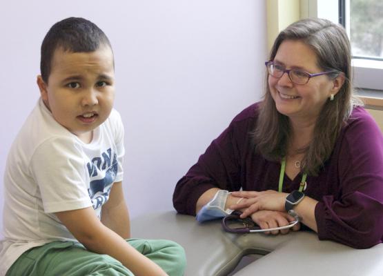 Boy with short dark hair and white t shirt sits on medical table with woman with long hair and glasses smiling at him