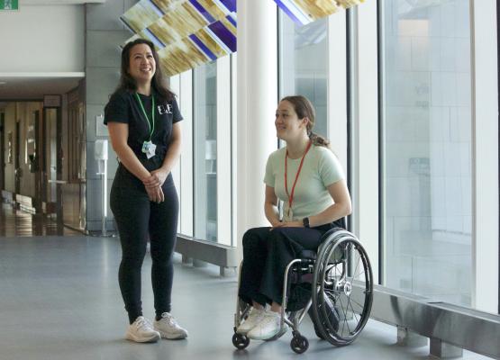 Two women smile beside floor to ceiling window, one in a wheelchair