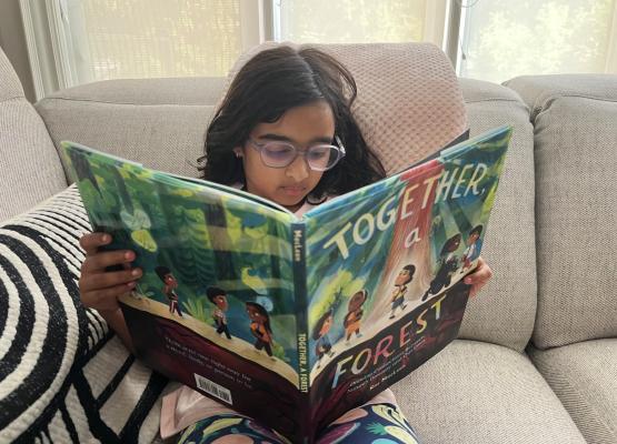 Girl with long dark hair and glasses reads large children's picture book