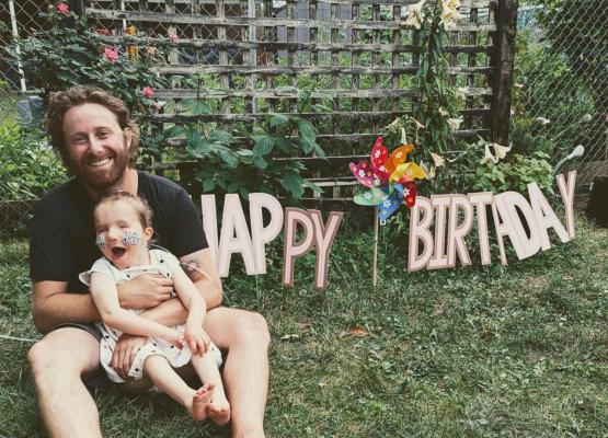 Man with beard holding young girl in lap with Happy Birthday sign behind