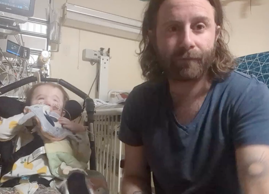 Man with beard beside child in stroller in hospital room with medical equipment
