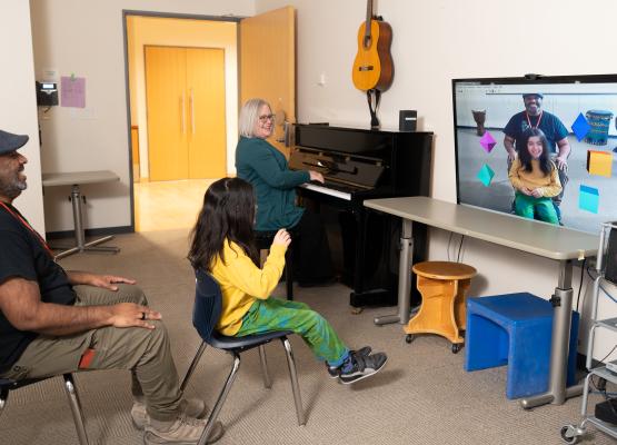 Child in yellow shirt sits in front of screen with icons with a woman on piano beside her and a man sitting behind her