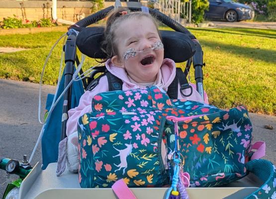 Young girl with oxygen prongs and huge smile sits in wheelchair with backpack on tray.