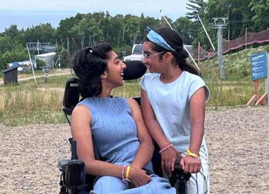 Teen girl in wheelchair and lavender outfit smiles at girl with bandana standing with arms on her chair