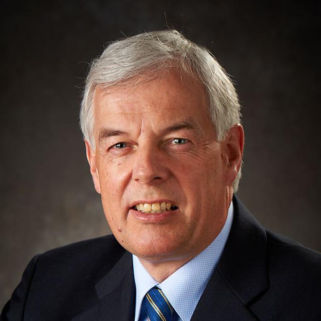 Man with gray hair wearing a dark blazer, light blue shirt, and a blue-striped tie
