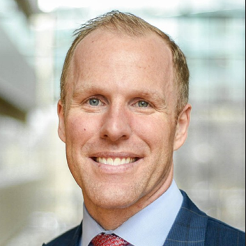 A smiling man with short blond hair and blue eyes. Wearing a dark blue plaid blazer, a light blue dress shirt, and a red patterned tie
