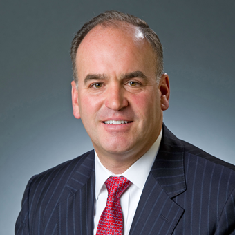 A smiling man with sort dark hair wearing a dark blue pinstriped blazer, white dress shirt, and red patterned tie