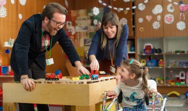 Man and woman laughing with child in walker over a play table 