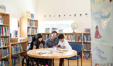 An adult sitting in the middle, to the left is a child on wheelchair and to the right is another child, in a library setting