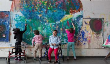 Four young boys with backs to the camera paining on a giant mural. Two of the children have walkers.