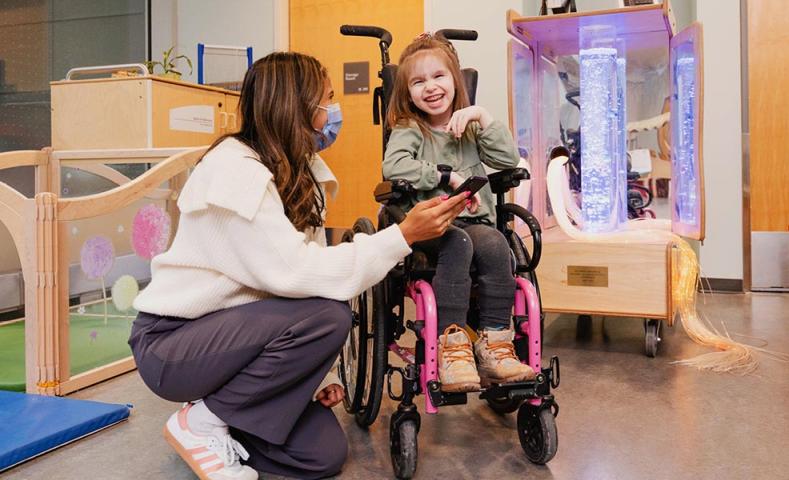 An adult with mask on, beside a child on a wheelchair
