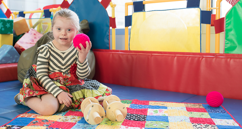 Little girl holding a red ball sitting on a quilt in a playroom
