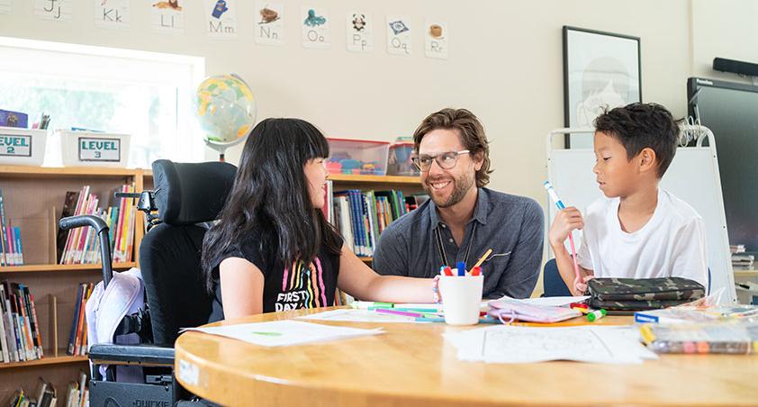 An adult is talking with a child who is on a wheelchair, and another child to the right and listening