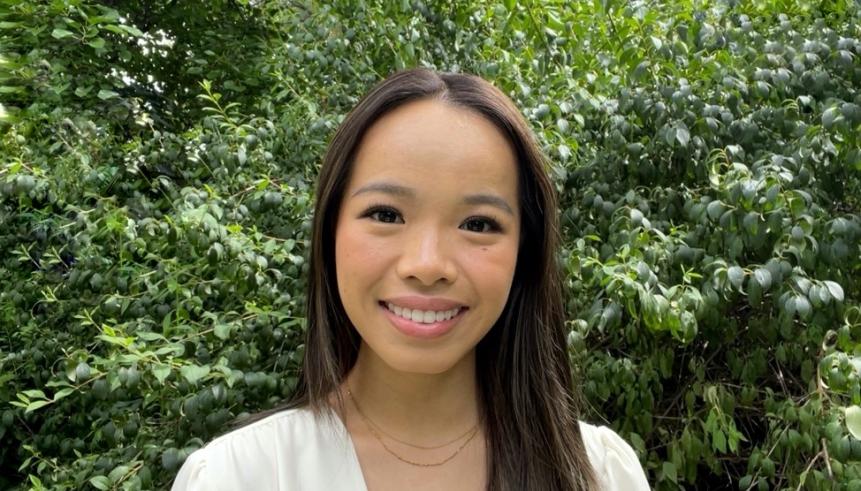 A woman with black hair wearing a cream coloured top. Behind them is greenery.