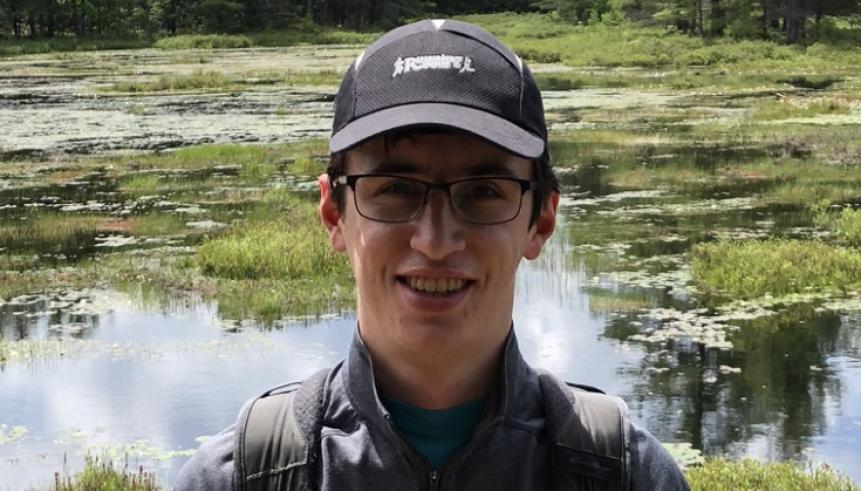 A man with a black shirt, with glasses and hat. Behind him, is greenery.